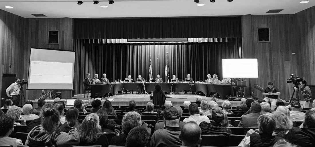 Black-and-white photograph of a packed high-school auditorium in Chatham, Virginia, where residents face the Pittsylvania County Board of Supervisors seated at a long table on the stage, April 15, 2025.