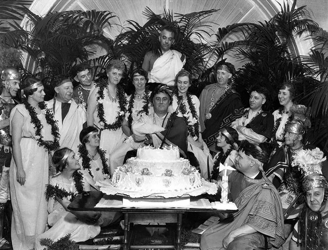 Black-and-white photograph of Franklin Roosevelt in a toga and laurel wreath, seated behind a tiered birthday cake among guests dressed as Romans: women in white robes and garlands, men in togas and helmets.